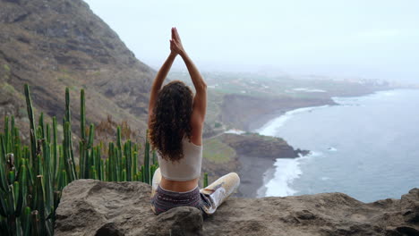 Amidst-mountainous-island-scenery,-a-young-woman-does-yoga,-seated-on-a-rock-atop-a-mountain,-meditating-in-Lotus-pose-while-gazing-at-the-ocean
